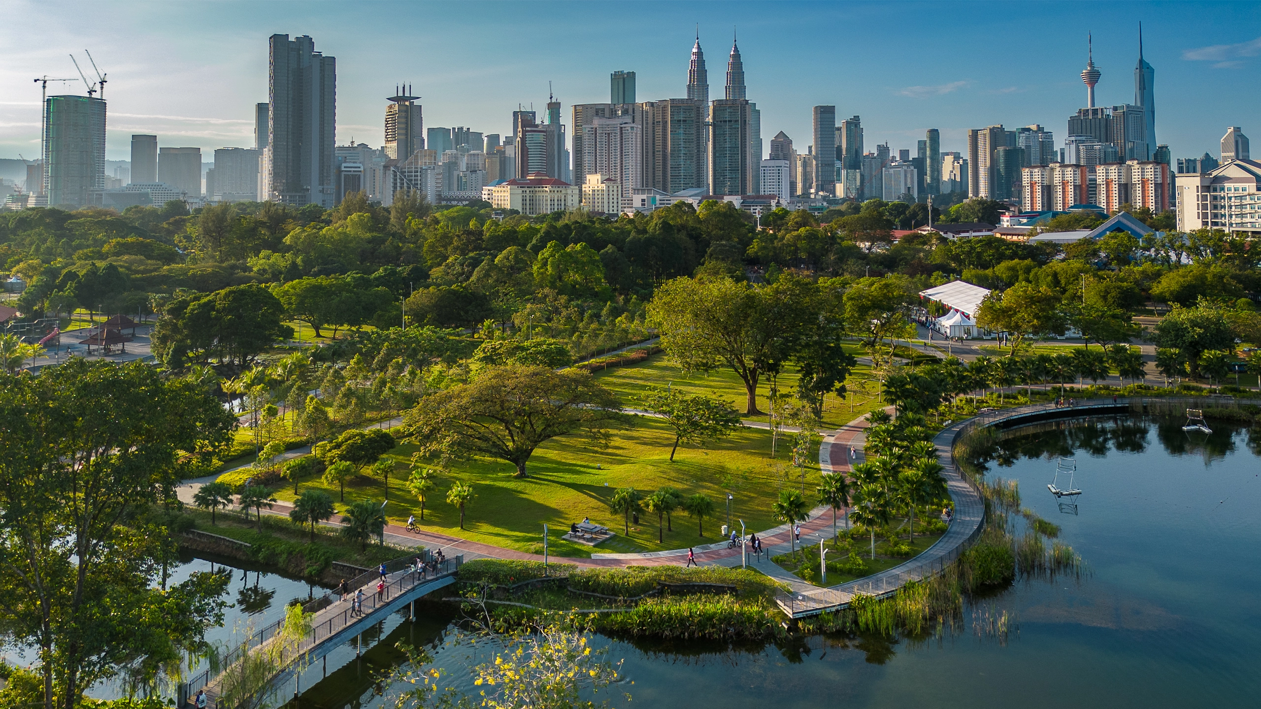 Aerial view of a city park with lush greenery, walking paths and a lake, set against a backdrop of tall sky scrappers